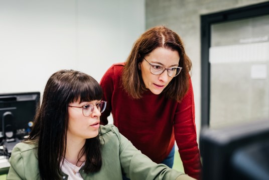 En kvinde siddende ved en computer og en kvinde stående bagved. Sammen kigger de på computerskærmen.